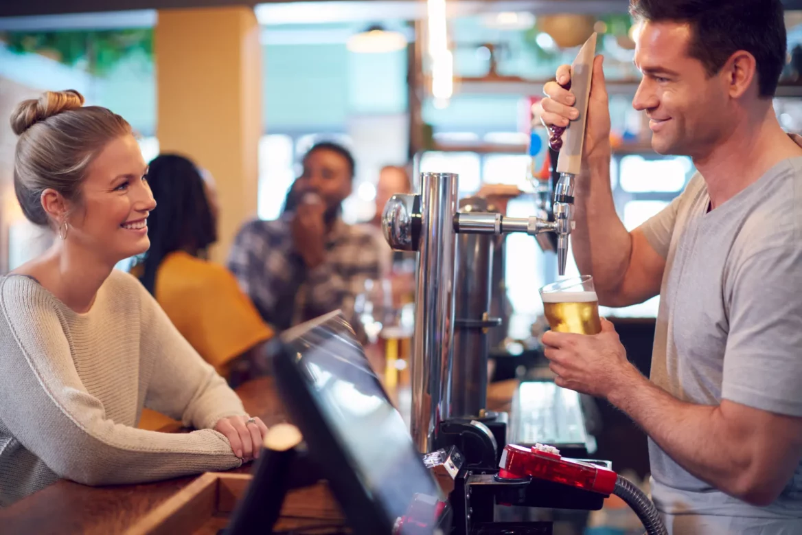 A bartender at a brewery pouring a beer for a woman customer after checking her age via ID